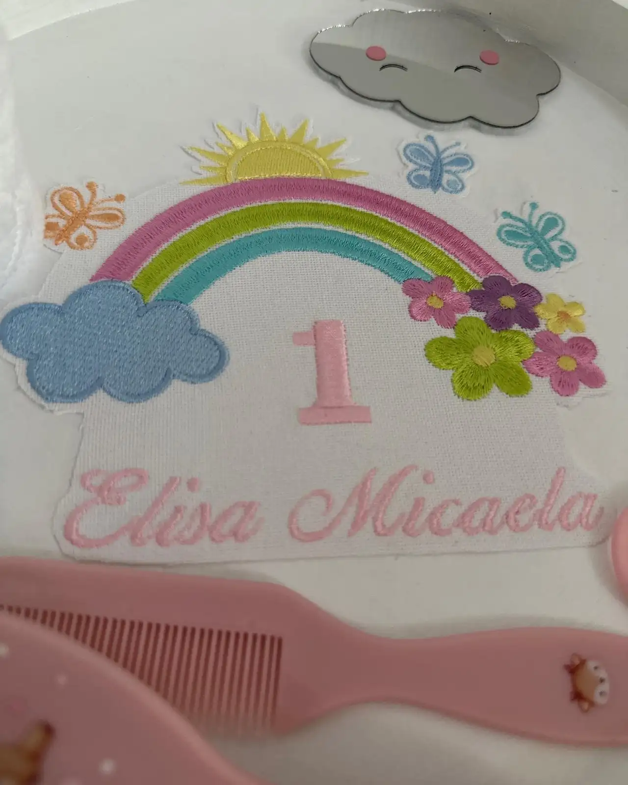 Prim-plan cu broderia colorată de curcubeu, nori, soare și fluturi de pe tăvița pentru moț